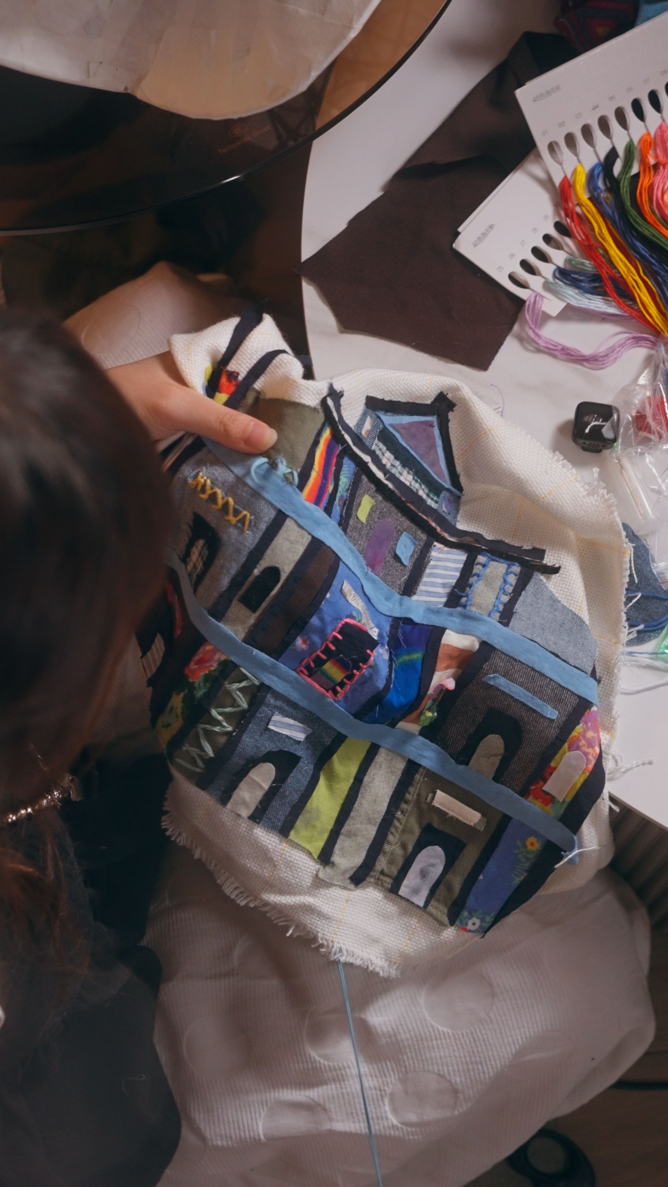 Detail of a textile artwork by Nicole Ng, featuring patchwork and embroidery techniques with colorful architectural motifs and layered fabric textures.